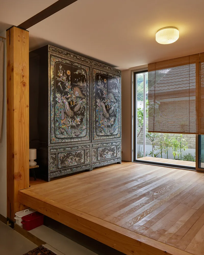 Timber-floored room with ornate lacquered cabinet against exposed column and glazed doors to courtyard with bamboo shade