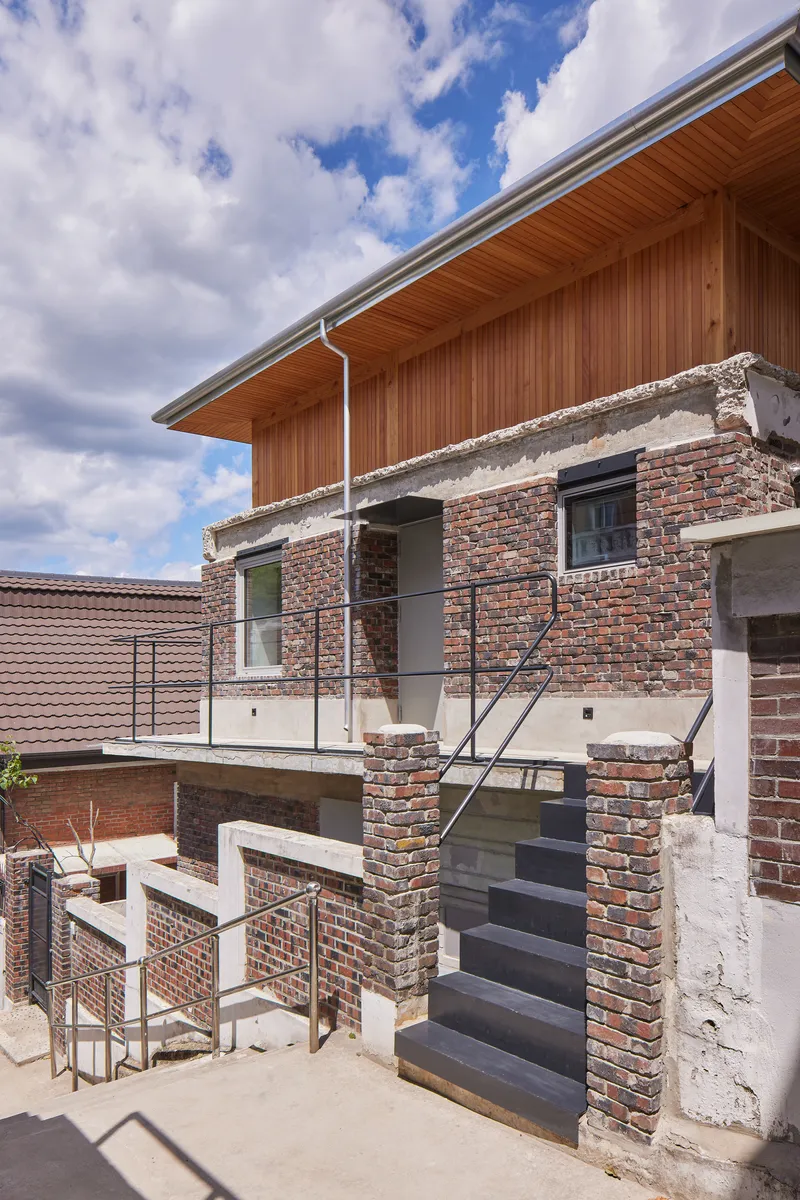 External staircase with dark treads rising between weathered brick columns beneath a timber-lined soffit and clouded sky