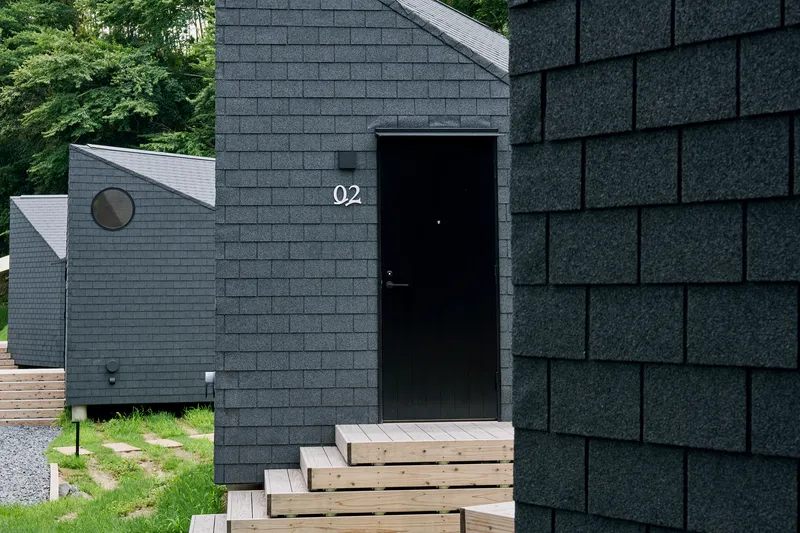 Black entry door with timber steps set in grey shingle-clad facade with round window visible