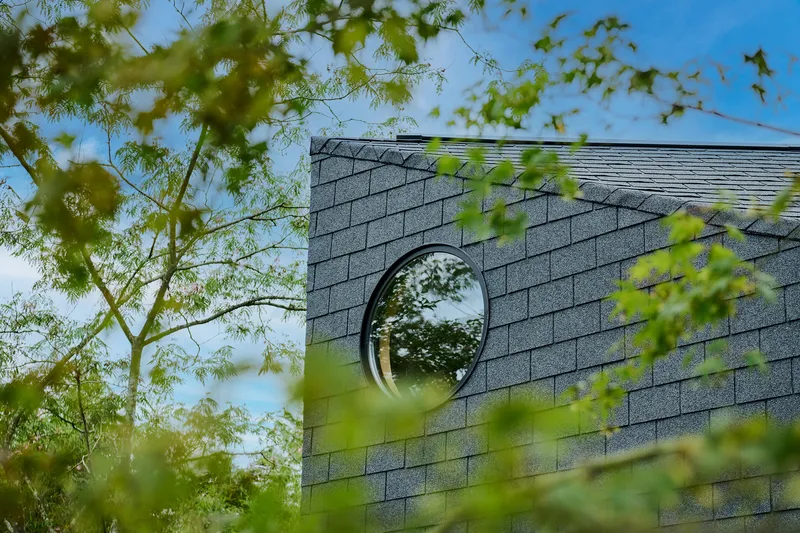 Circular window in grey shingled wall framed by spring leaves and blue sky
