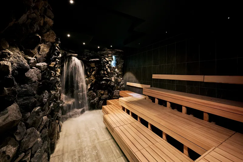 Timber sauna benches beside rock waterfall illuminated at night with dark tiled wall