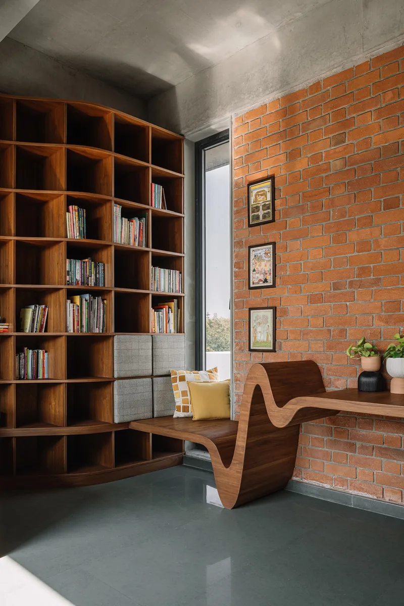 Reading nook with floor-to-ceiling timber bookshelf against red brick wall and curved desk beneath window