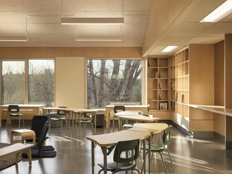 Classroom with plywood ceiling, built-in timber shelving, and tables facing woodland views through floor-to-ceiling windows