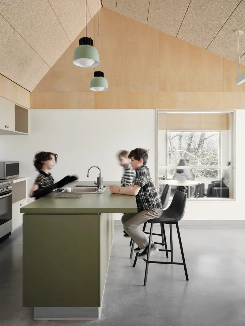 Kitchen island with olive-green countertop beneath plywood vaulted ceiling and pendant lights, children in motion