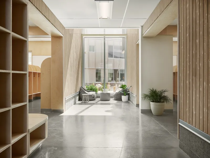 Corridor with timber-clad walls and built-in plywood shelving leading to glazed courtyard view with potted plants