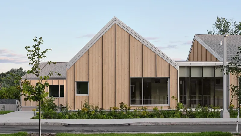 Street view of vertical timber-clad gabled volumes with planted beds and young trees