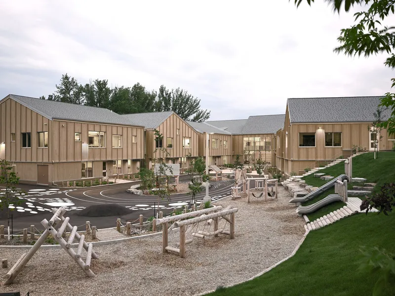 Courtyard view showing timber-clad wings enclosing a gravel playground with wooden climbing equipment at dusk