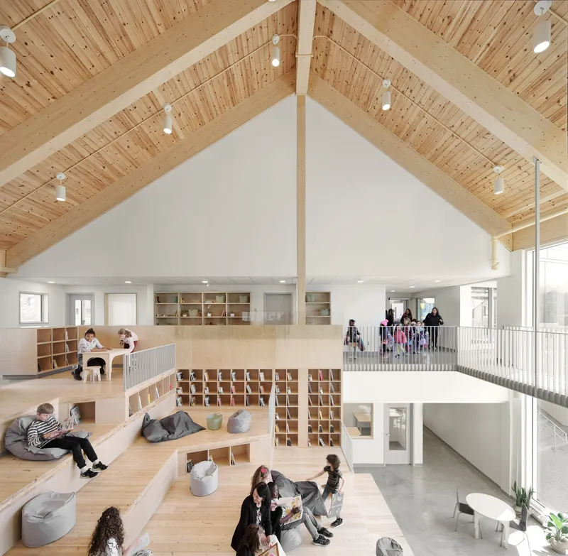 Double-height interior with exposed timber ceiling, plywood shelving, and children occupying tiered seating platforms