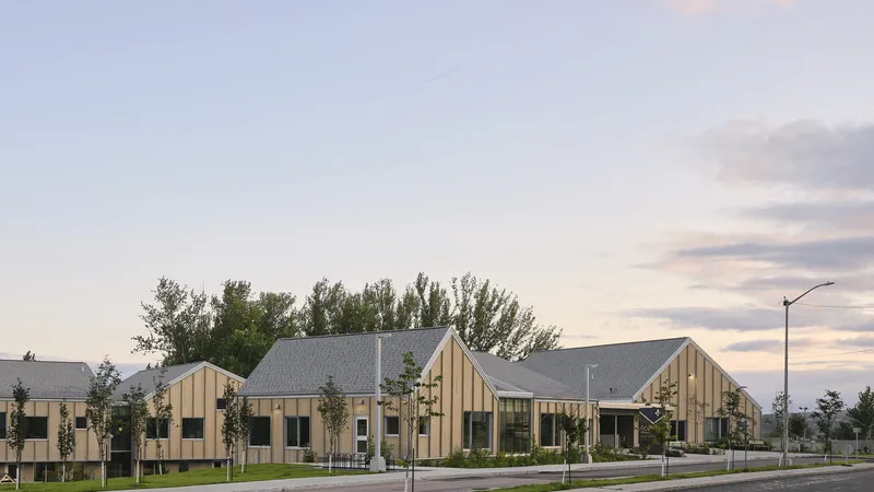 Street view of timber-clad gabled volumes with young trees in a landscaped lawn at sunset