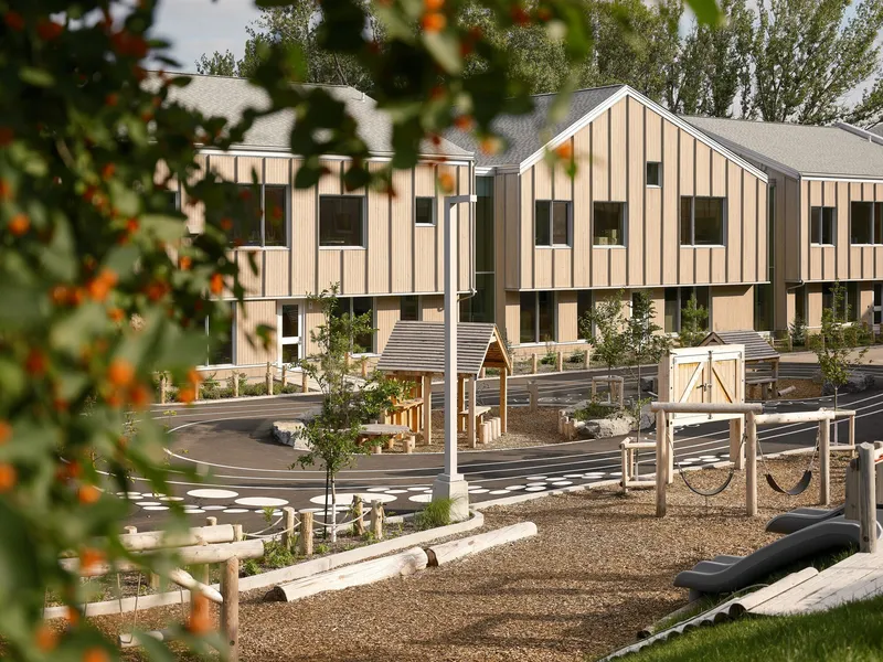 Gabled timber volumes around a play yard with climbing equipment seen through autumn foliage