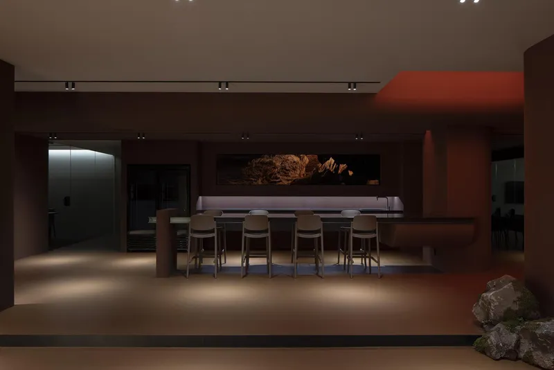 Dining area with long table and upholstered stools beneath brown walls and recessed ceiling lighting