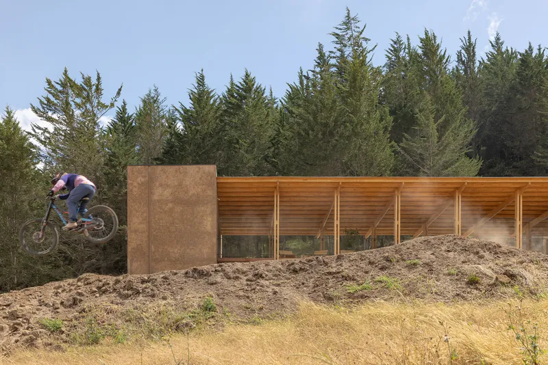 Cyclist jumping earthen berm beside rammed earth wall and timber-framed glazed facade in afternoon sun