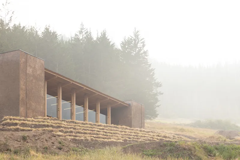 Timber-clad volume with exposed columns and glazing set into a hillside under dense fog