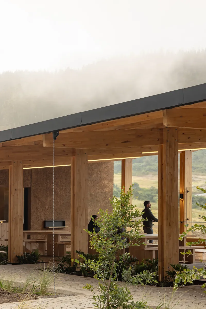 Interior view through timber columns toward two figures and planted beds with misty hills beyond