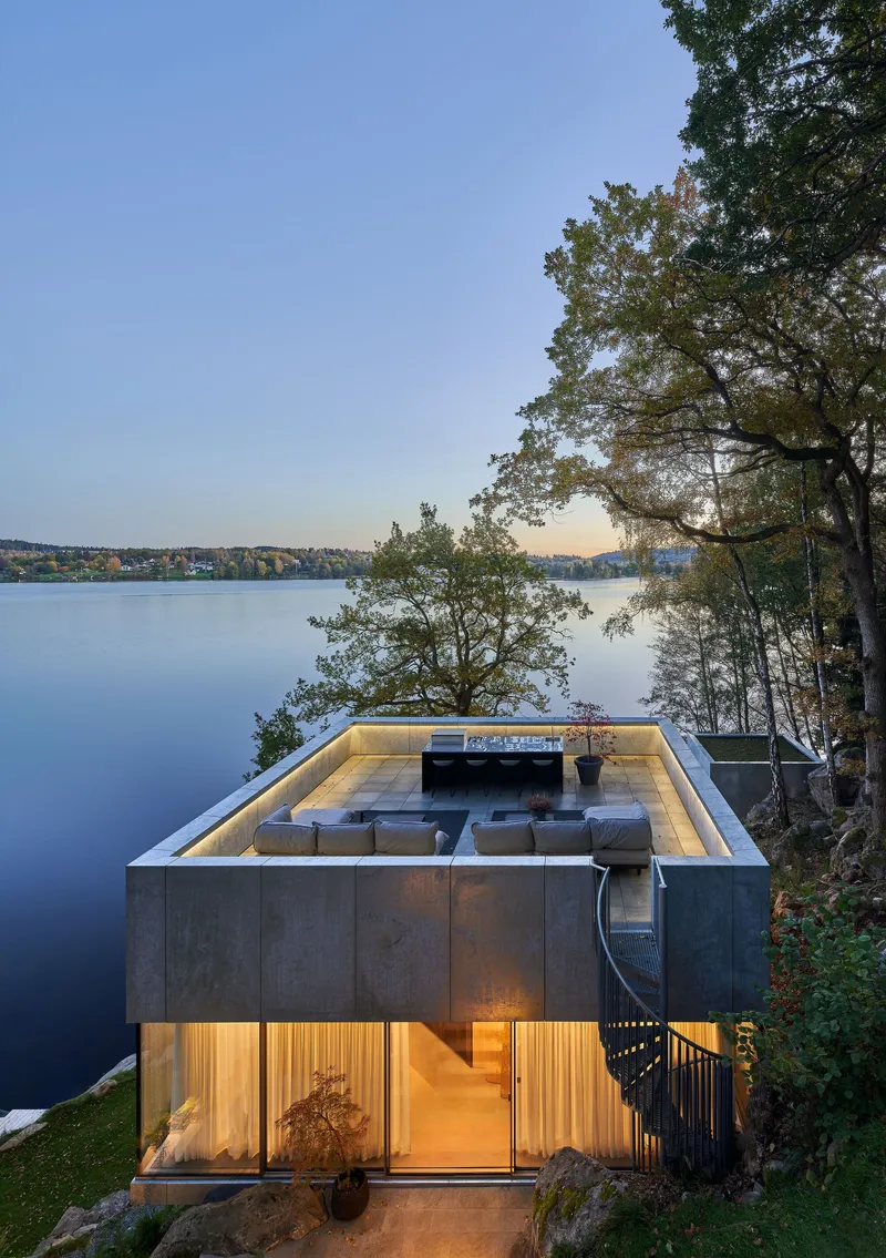 Lakeside facade at dusk with roof terrace above illuminated lower level timber and glass walls