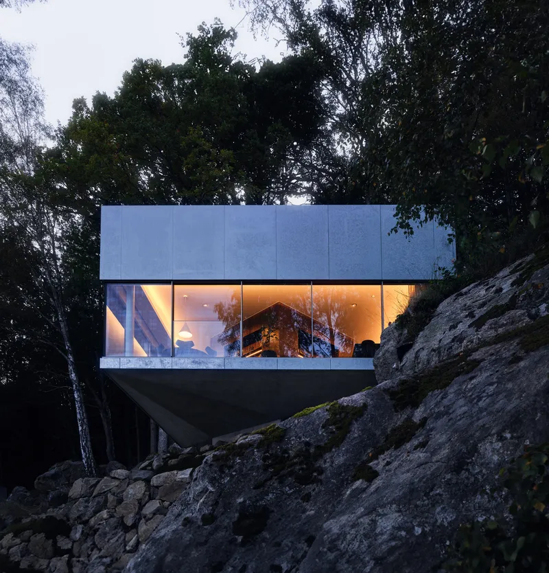 Cantilevered glass-walled volume with illuminated interior perched above a rocky slope at dusk