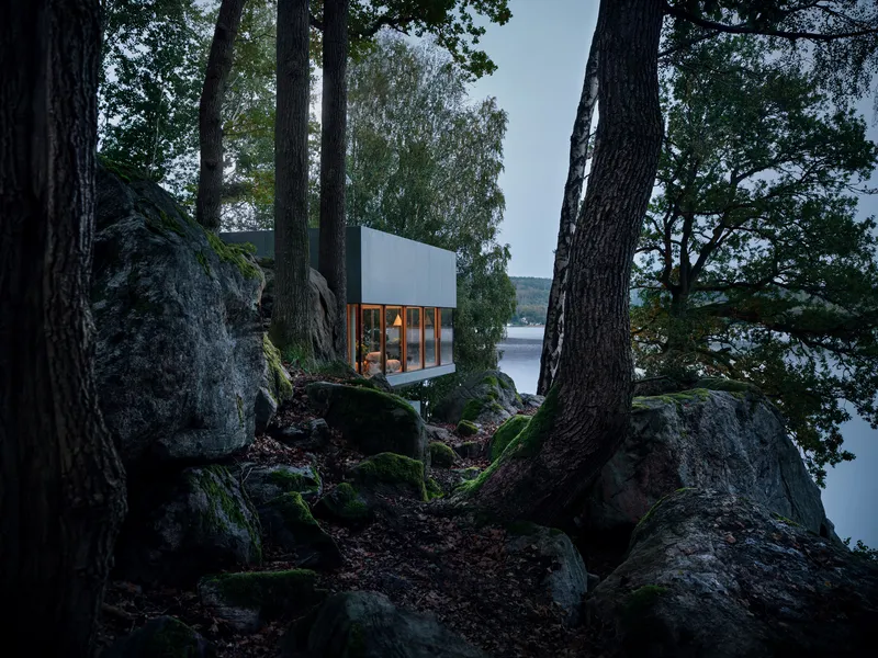 White cubic volume with glazed facade glimpsed through moss-covered boulders and tree trunks beside a lake