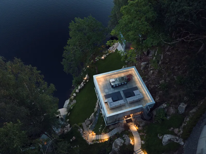 Aerial view of flat-roofed glass pavilion with terrace seating nestled among boulders beside dark water