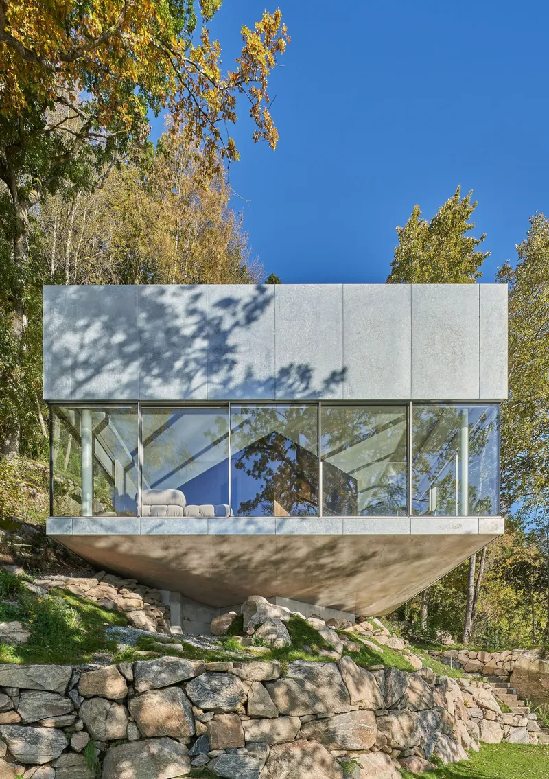 Glazed volume cantilevered on sloped concrete supports above a stone retaining wall with dappled tree shadows