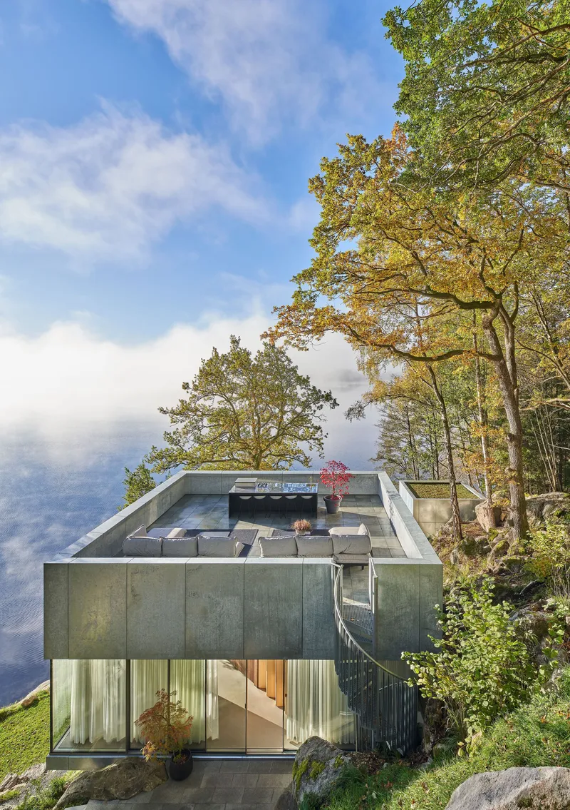 Two-story concrete volume with glazed lower level and rooftop terrace perched on a rocky slope among autumn trees