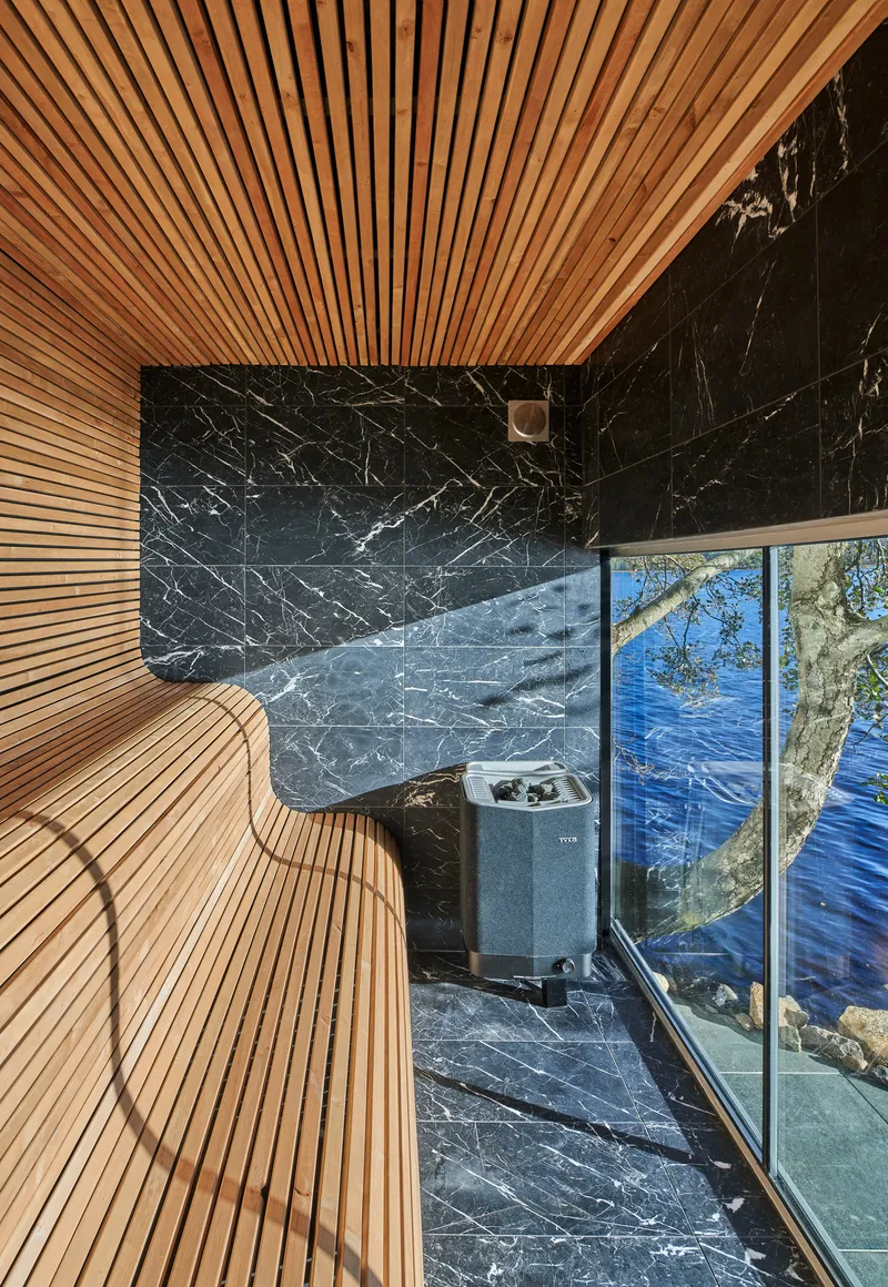 Sauna with slatted timber ceiling and benches alongside black veined marble wall facing the water