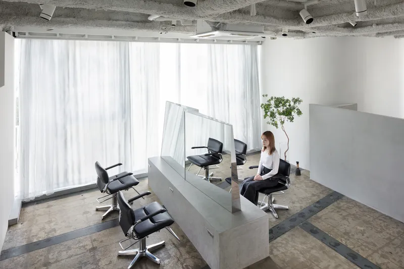 Hair salon interior with concrete workstations, glass dividers, and sheer white curtains diffusing daylight