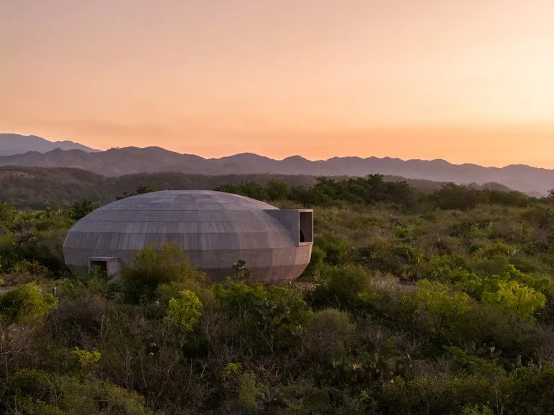 Timber-clad elliptical volume with rectangular apertures amid native brush at sunset with distant mountain range