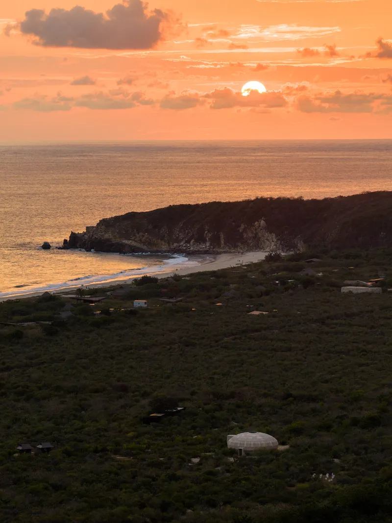 Aerial view of dome structure in dense vegetation with coastline and setting sun over water