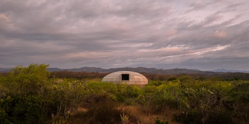 Domed structure with timber cladding and rectangular opening set among desert scrub under overcast skies