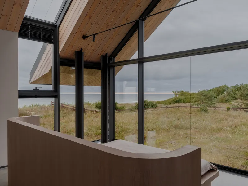 Interior corner with curved timber joinery and floor-to-ceiling black-framed glazing overlooking grassy dunes