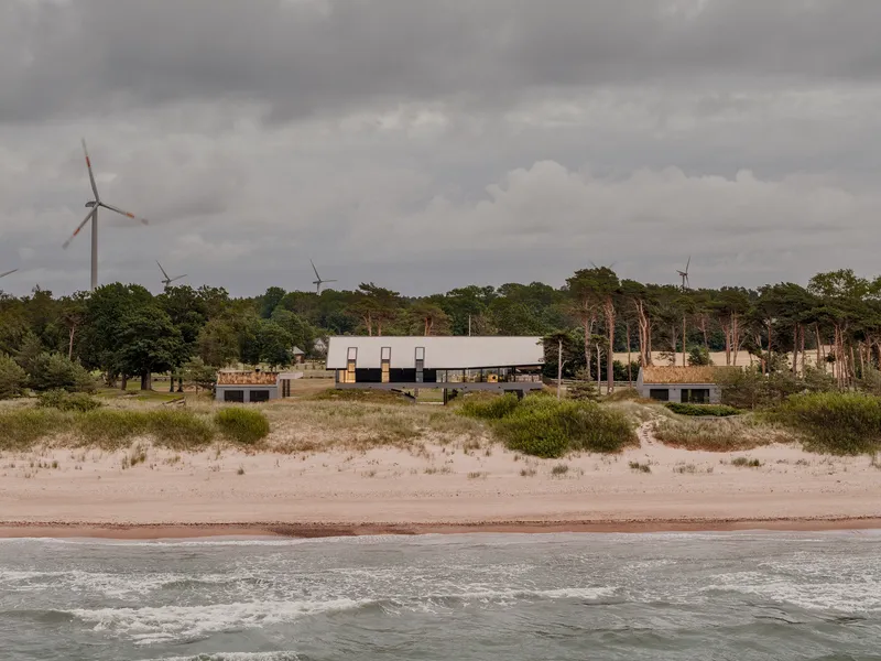 View from the beach showing the low-slung volumes with metal roof amid dune grasses and distant wind turbines