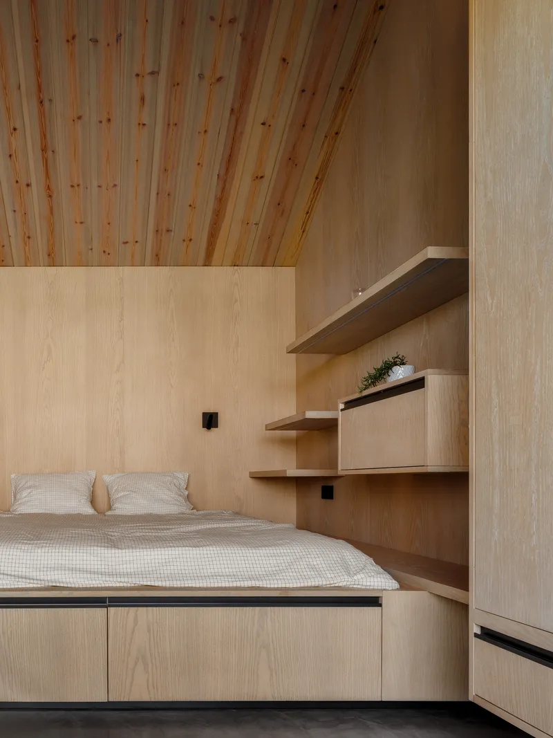 Bedroom with plywood wall panels, floating shelves, and sloped timber ceiling above the bed