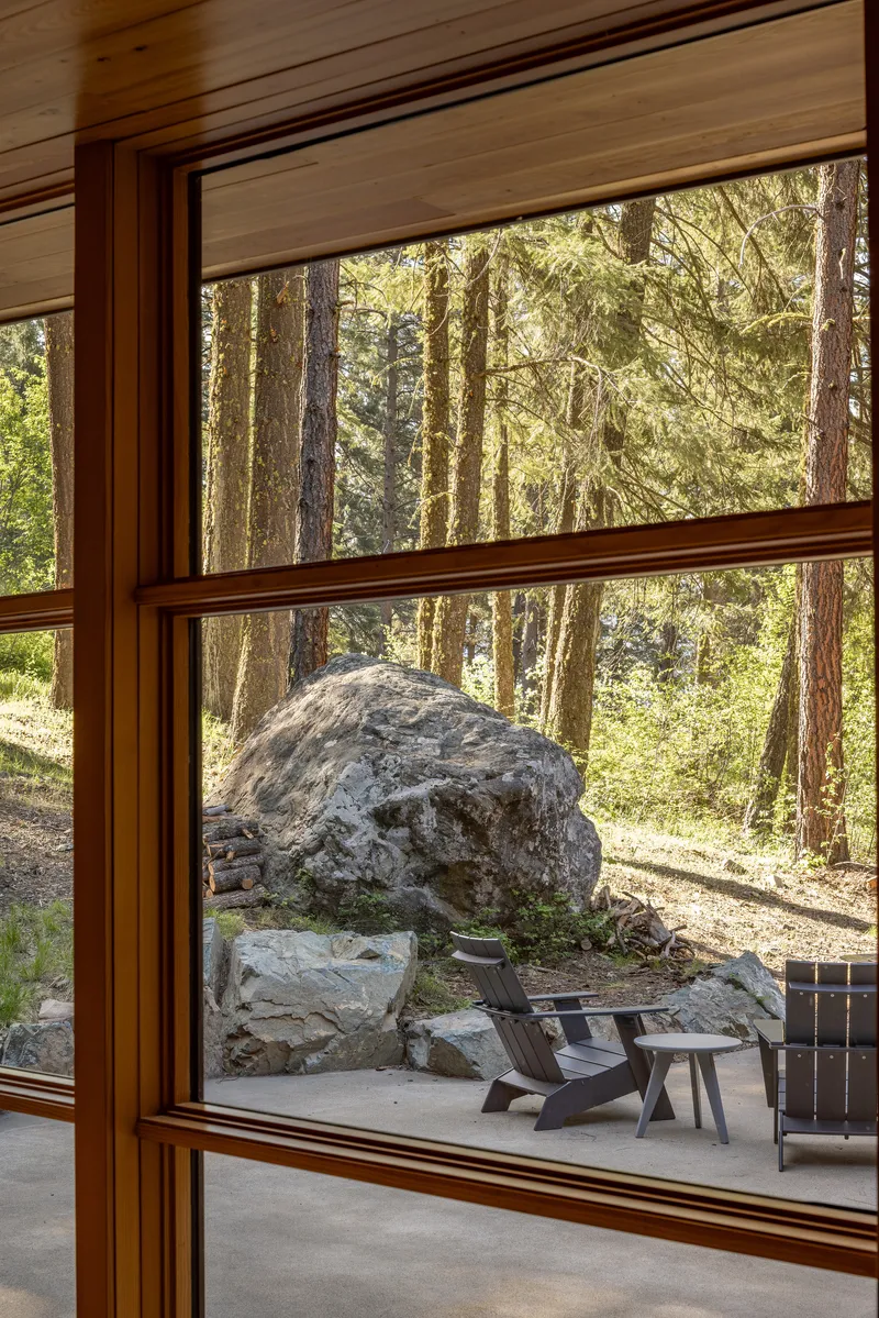 Timber-framed window looking out to a concrete patio with a large boulder and conifer trees beyond