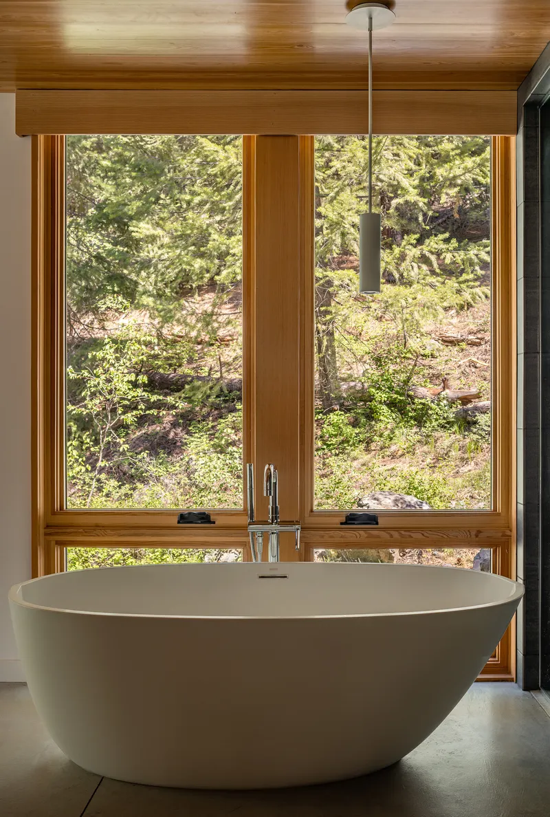 Freestanding bathtub positioned beneath a timber-framed corner window overlooking a wooded hillside