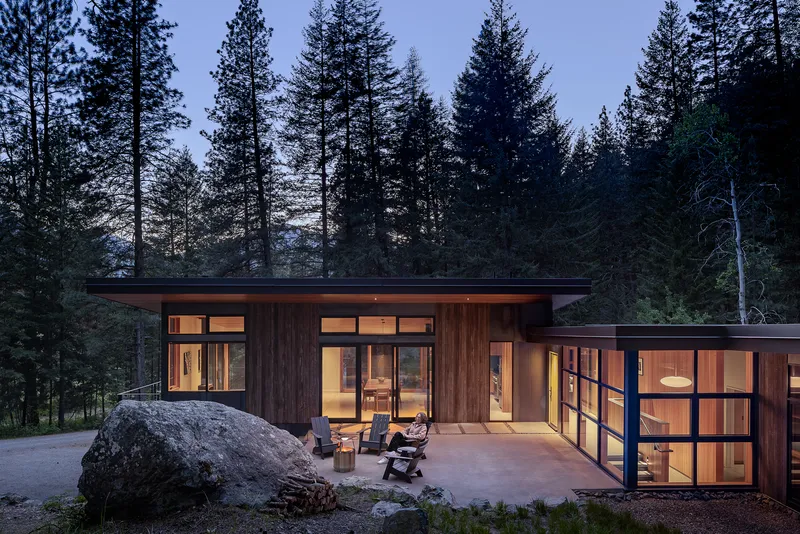 Timber-clad volumes with flat roofs and glazed walls glowing at dusk among tall conifers