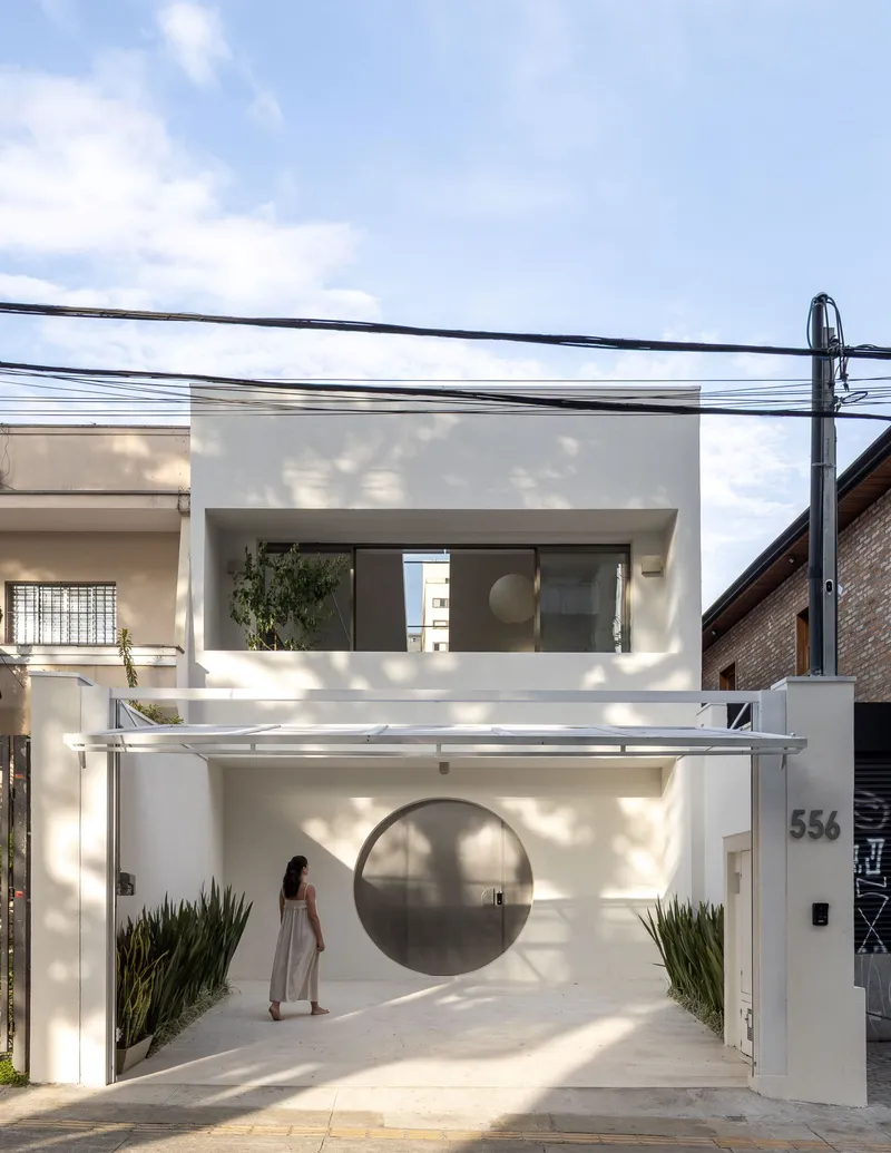 Street facade with white stucco walls, circular entry portal and a figure walking through dappled sunlight