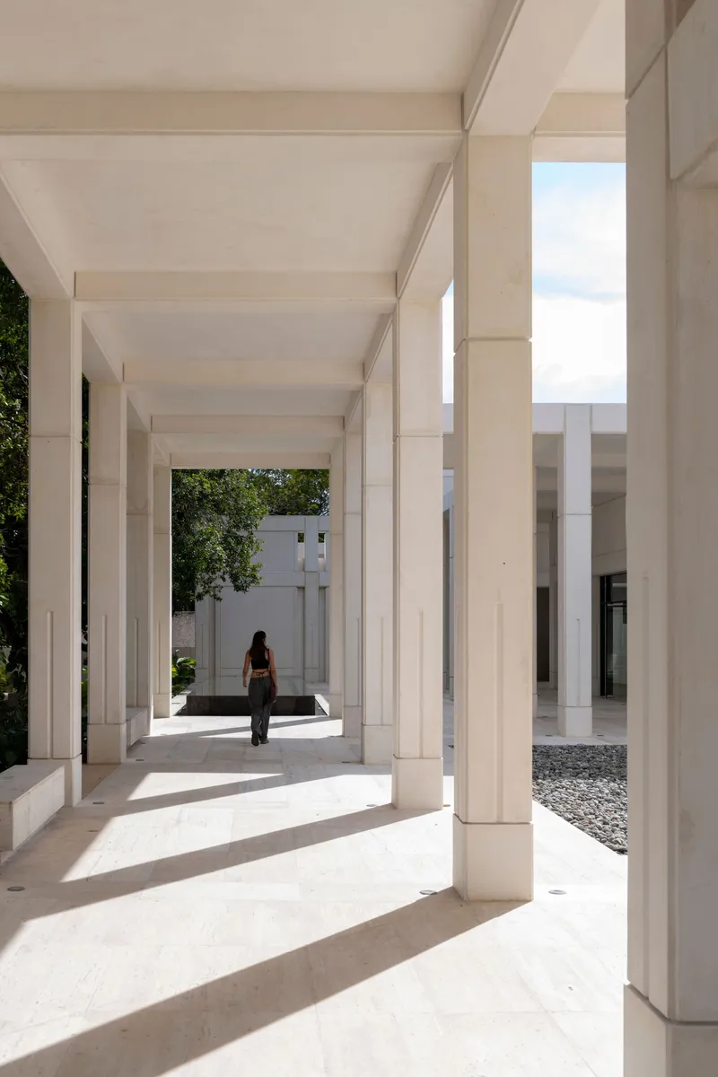 Covered walkway with coffered ceiling and white columns casting sharp shadows, a figure walking toward a courtyard
