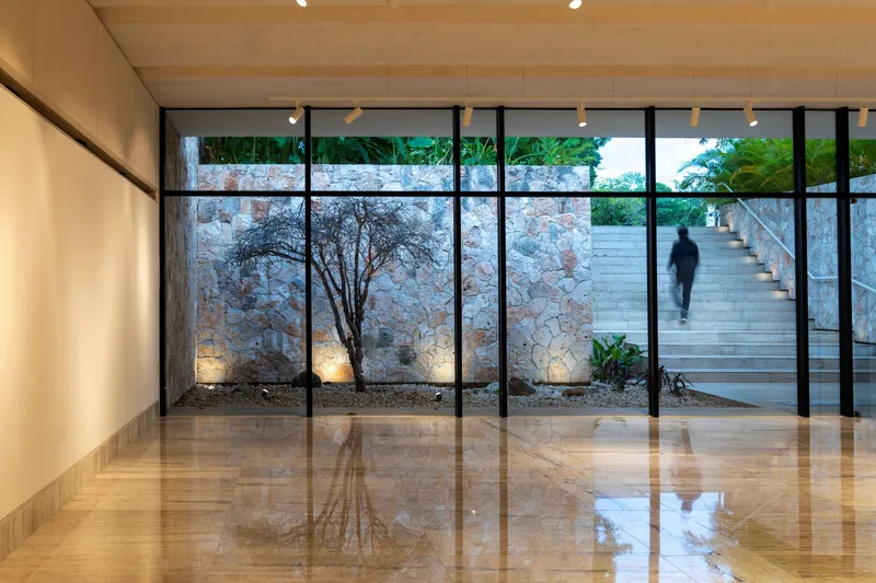 Interior gallery with polished stone floor and glazed wall revealing lit courtyard tree and exterior stair