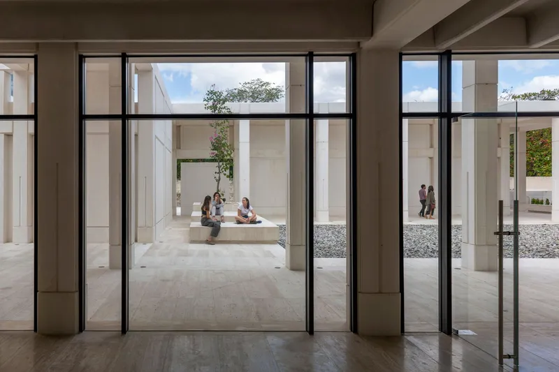 View through steel-framed glass doors into courtyard where visitors gather on concrete benches among gravel