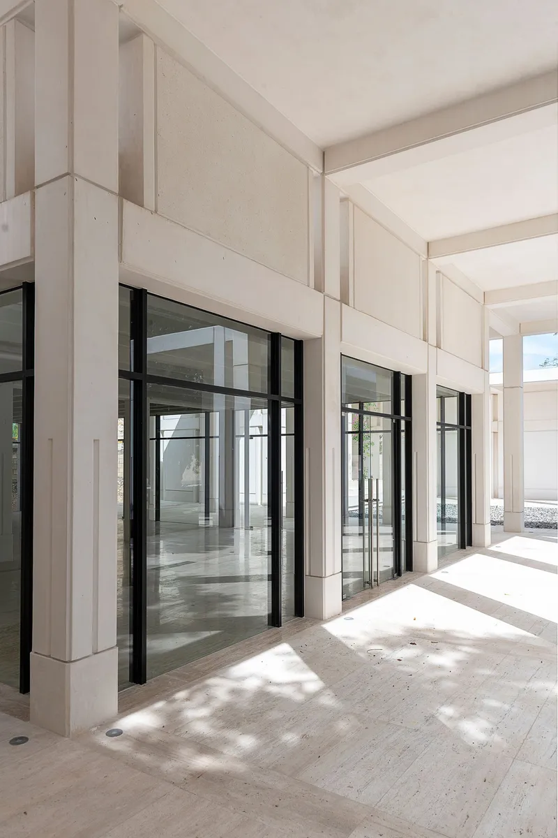 Covered portico with pale stone columns and black-framed glass doors casting dappled shadows on the floor