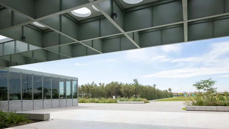 Covered terrace with green steel canopy and skylights overlooking a concrete plaza and planted landscape