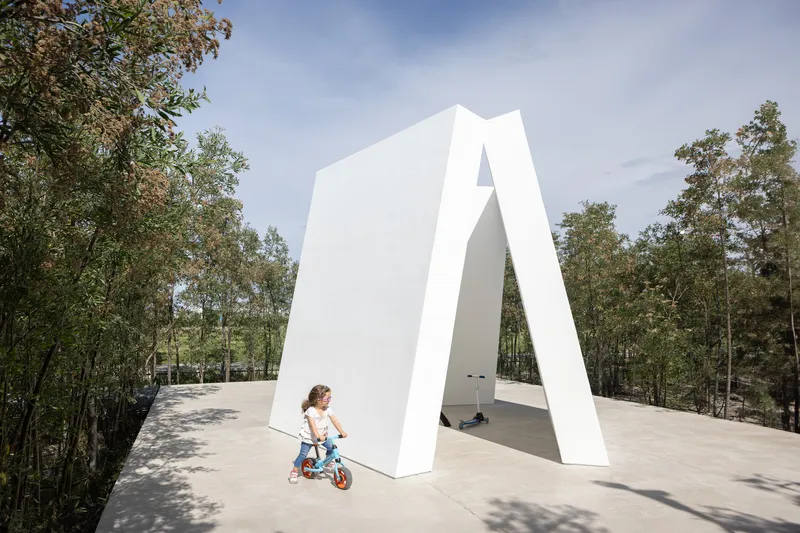 White angular portal structure framing a view through to woodland with child on bicycle