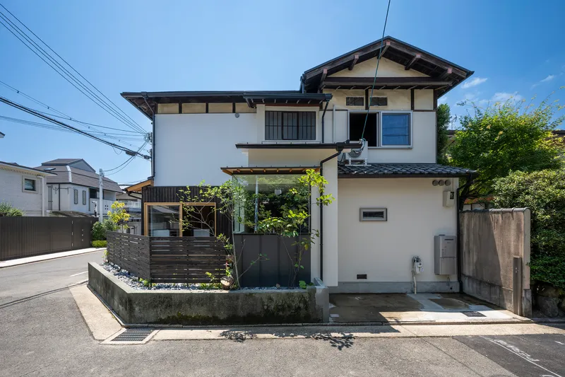 Street facade with white stucco walls, dark horizontal screens and small planted garden on a clear day