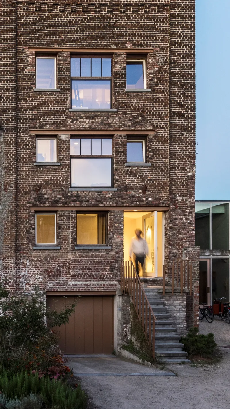 Facade at dusk with reclaimed brick wall and illuminated entry doorway at top of stone stair