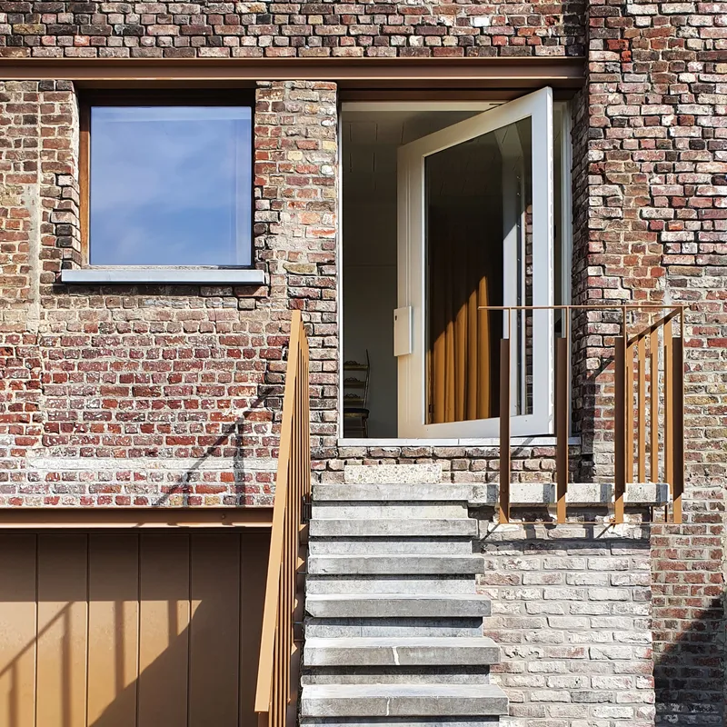 Outdoor stair with timber handrail ascending to glazed doorway framed by weathered brick