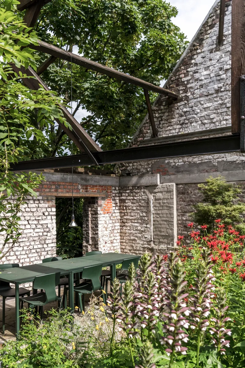 Garden dining area with steel table under painted brick walls and angled timber beams among flowering plants