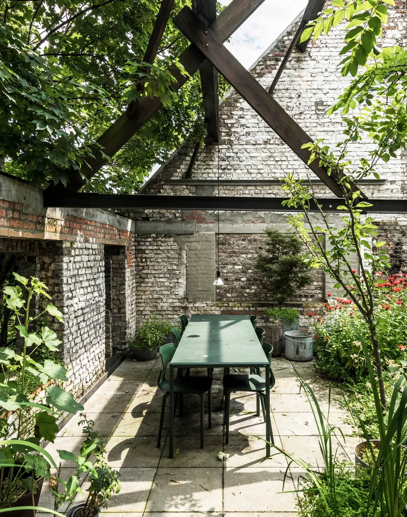 Outdoor dining table under timber roof frame with whitewashed brick gable wall and dappled tree shade