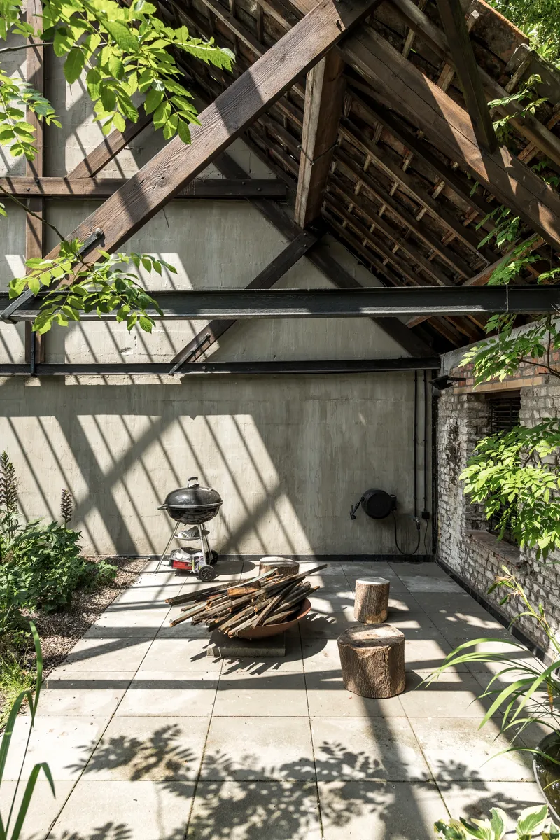 Paved courtyard with charcoal grill and firewood beneath angled timber structure casting diagonal shadows