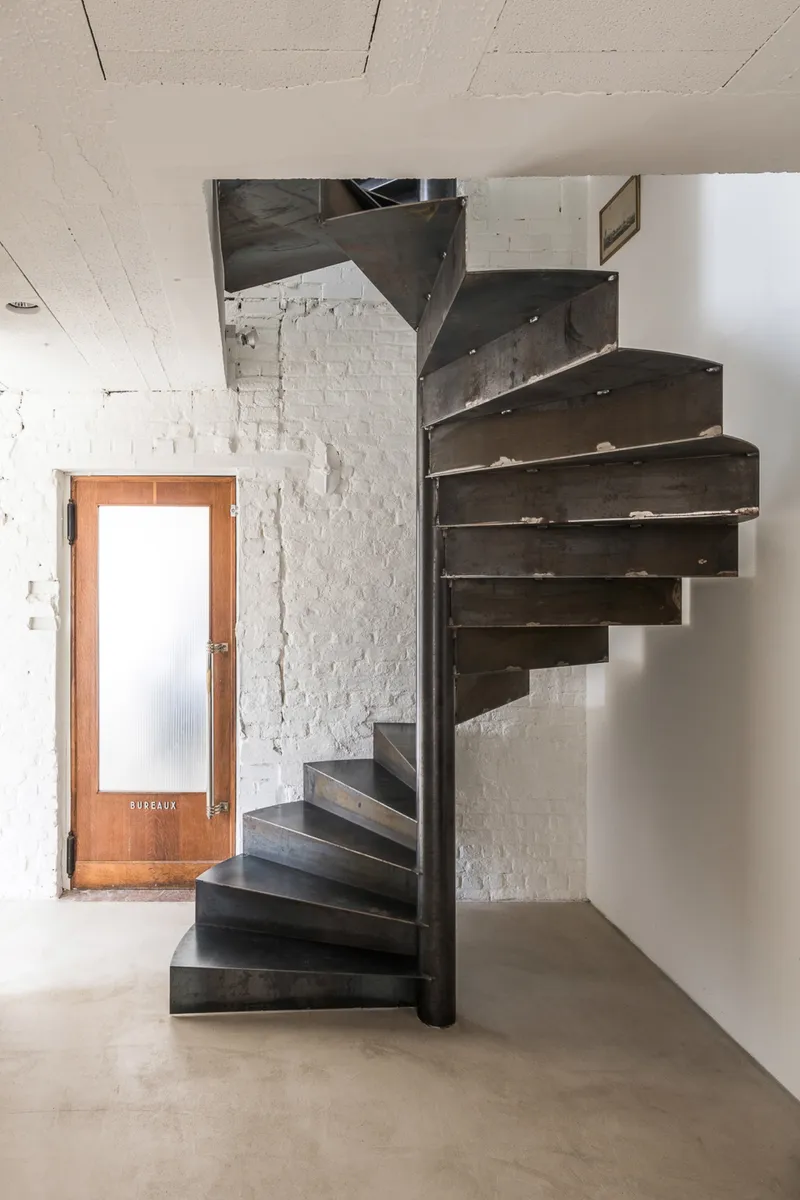 Black steel spiral staircase in a white-walled interior with glazed entrance door and polished concrete floor