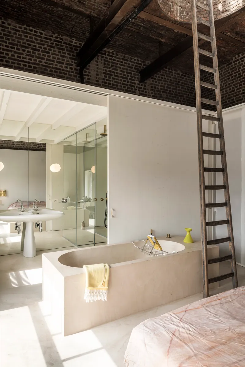 Bathroom with sunken concrete tub, glass shower enclosure, and narrow ladder ascending to exposed brick vault